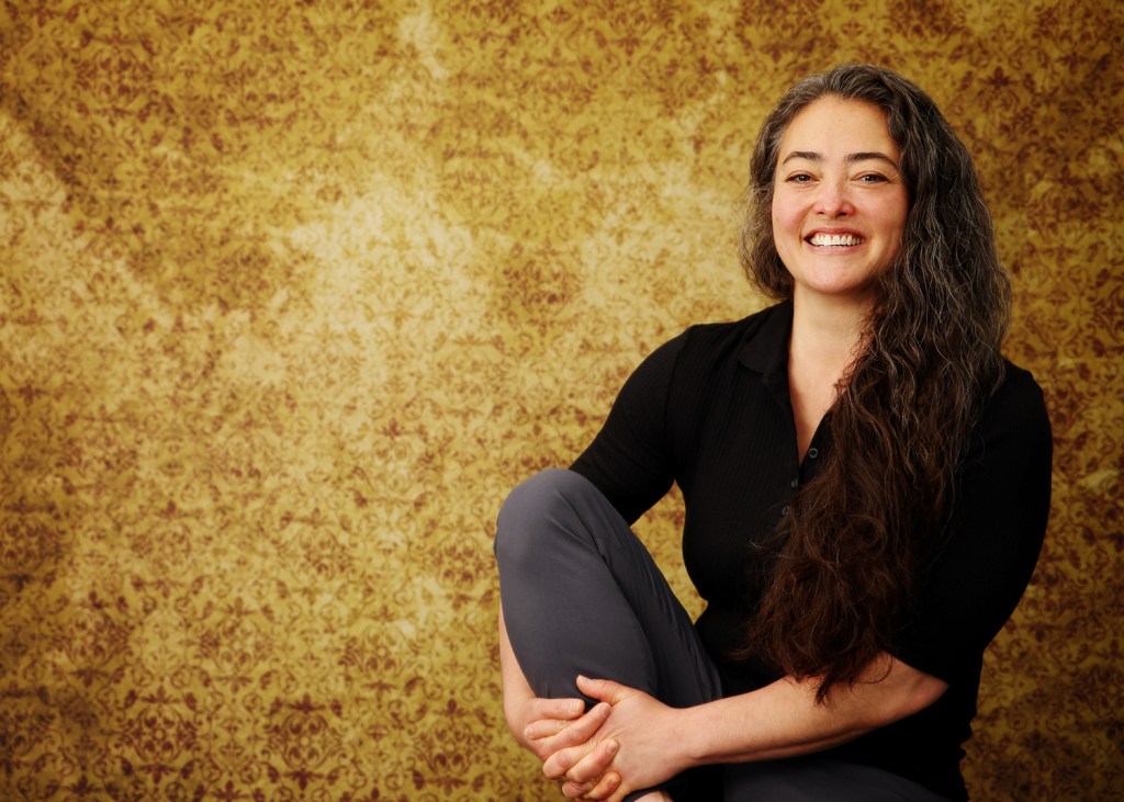Woman sitting, holding one knee, wearing black shirt and gray pants with an yellow background